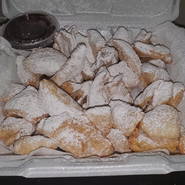 A white takeout container filled with powdered sugar-dusted beignets and a side of chocolate dipping sauce.