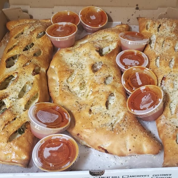 Several golden-brown calzones with visible cheese and herbs, served with small cups of marinara sauce in a box.